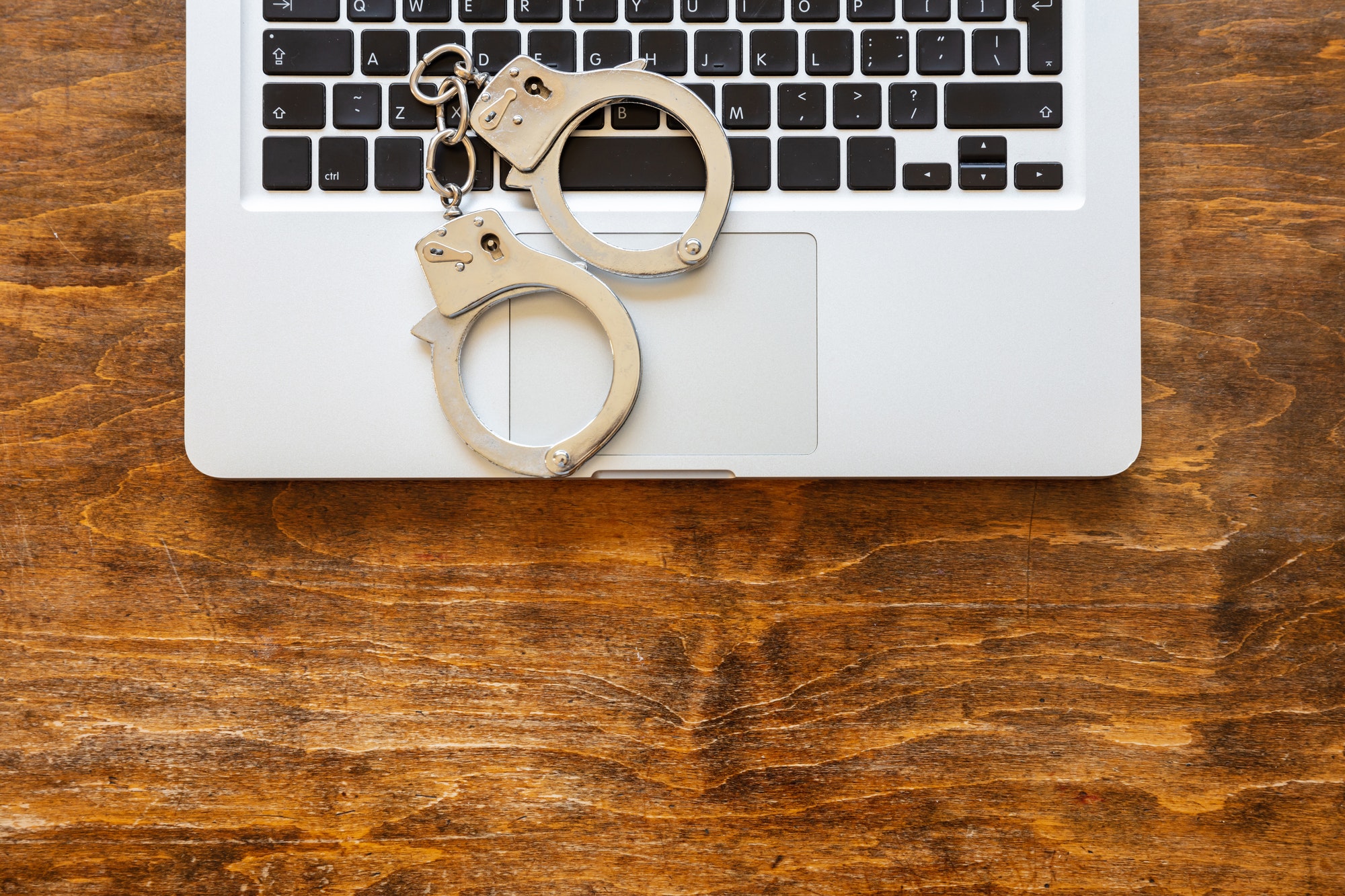 Handcuffs on a laptop, wooden office desk background, top view.