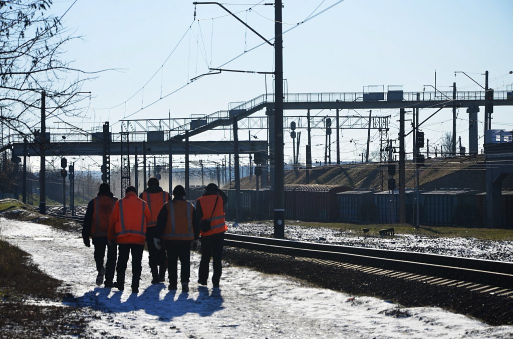 Several railway workers in signaling dirty orange uniforms are on the road next to the railway line