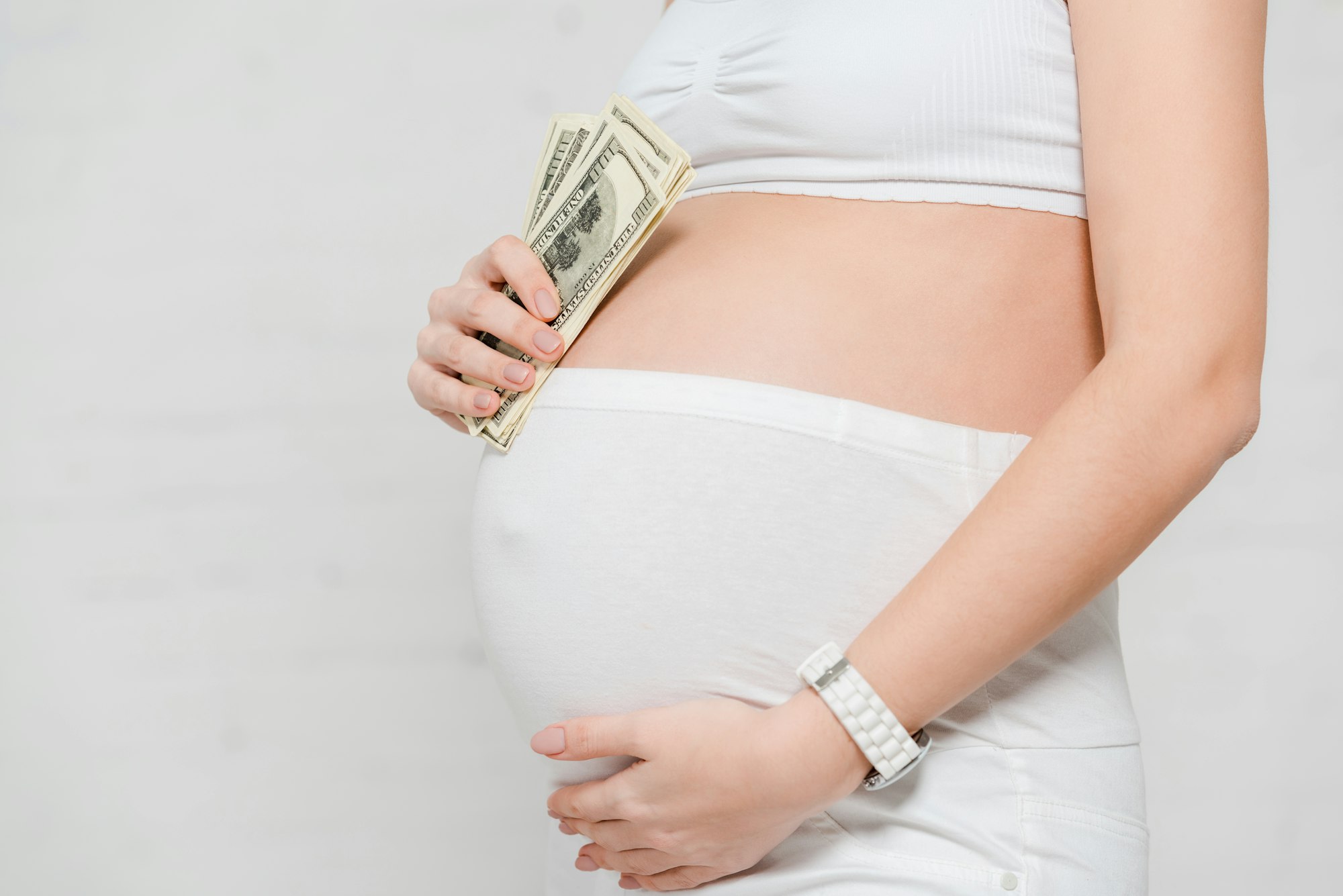 Cropped view of pregnant woman holding dollar banknotes and touching belly on grey background,