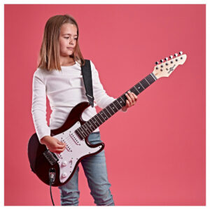 A girl playing the Gear4music 3/4 LA Electric Guitar with a strap and cable plugged in, stood in front of a pink background