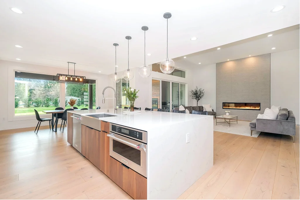 Contemporary kitchen featuring a big island with an oven and sink, with living room in the background and big window leading to garden on the left.