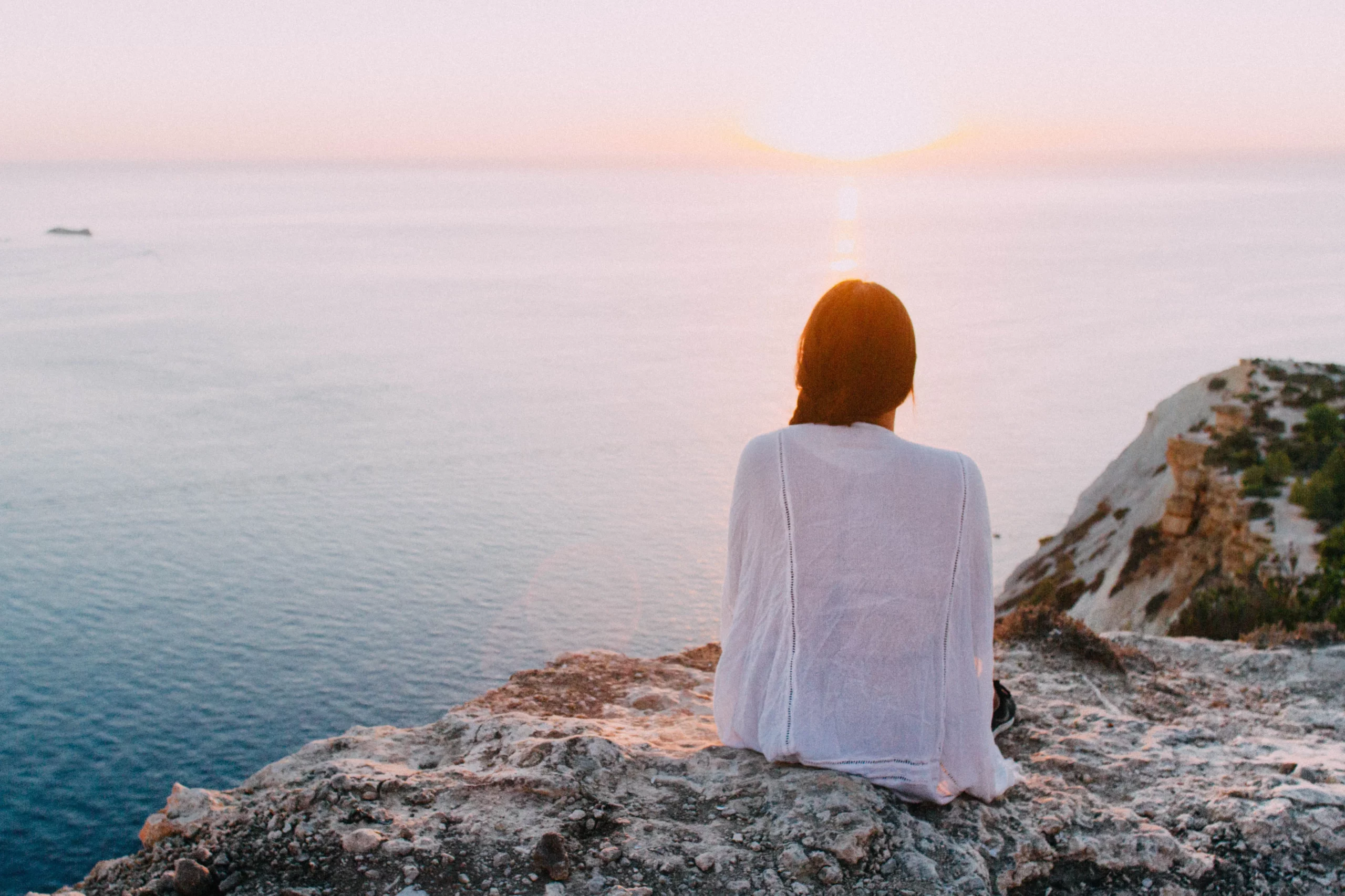 Girl looking into the sea from a rockly cliff at sunset time.