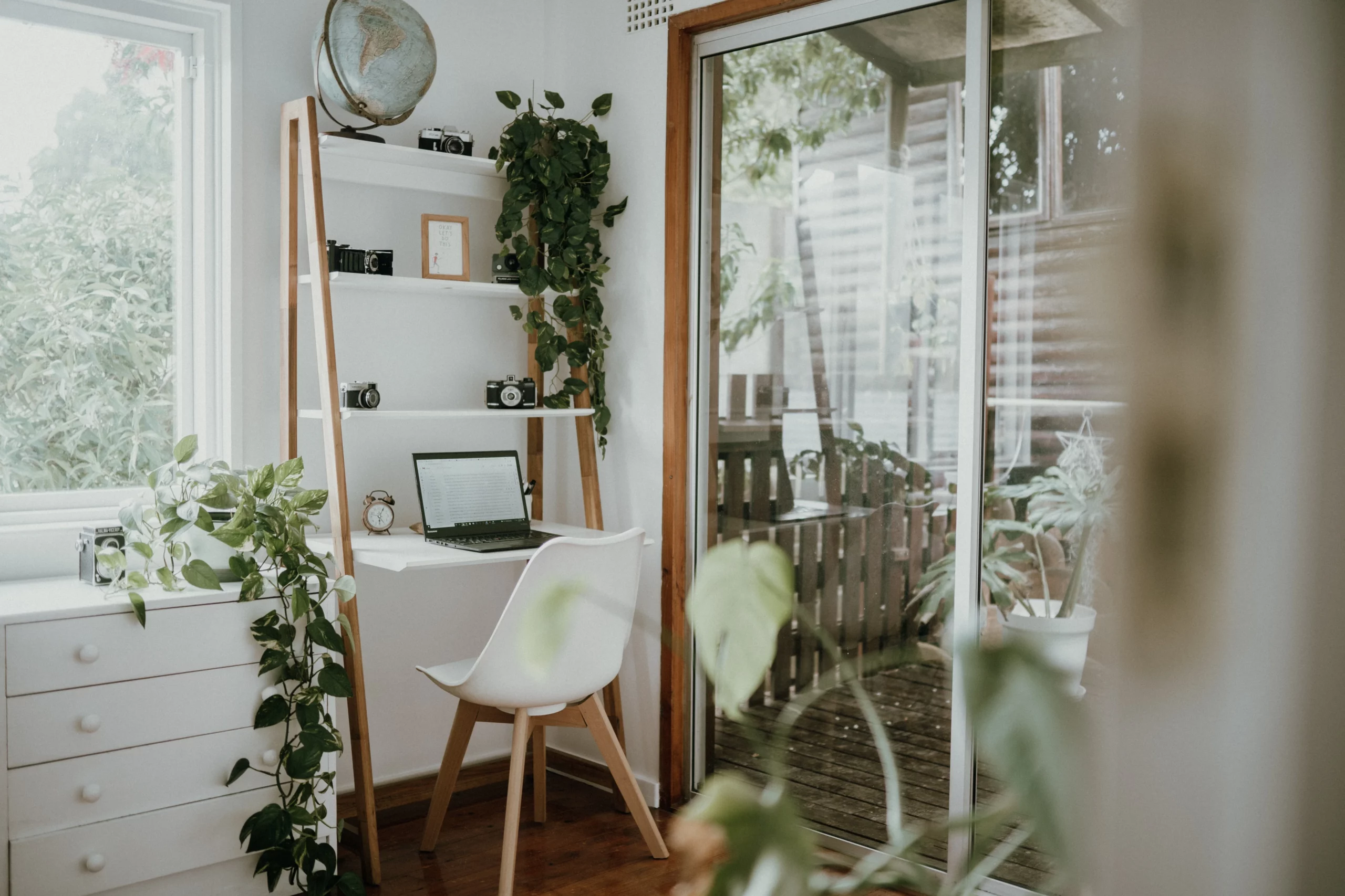 Light and airy remote work area surrounded by while furniture and green plants. Desk sits under a wall shelf area besides a big window that looks onto a green garden. Under the window, there is a large while dresser. There is a sun room to the right of the desk.