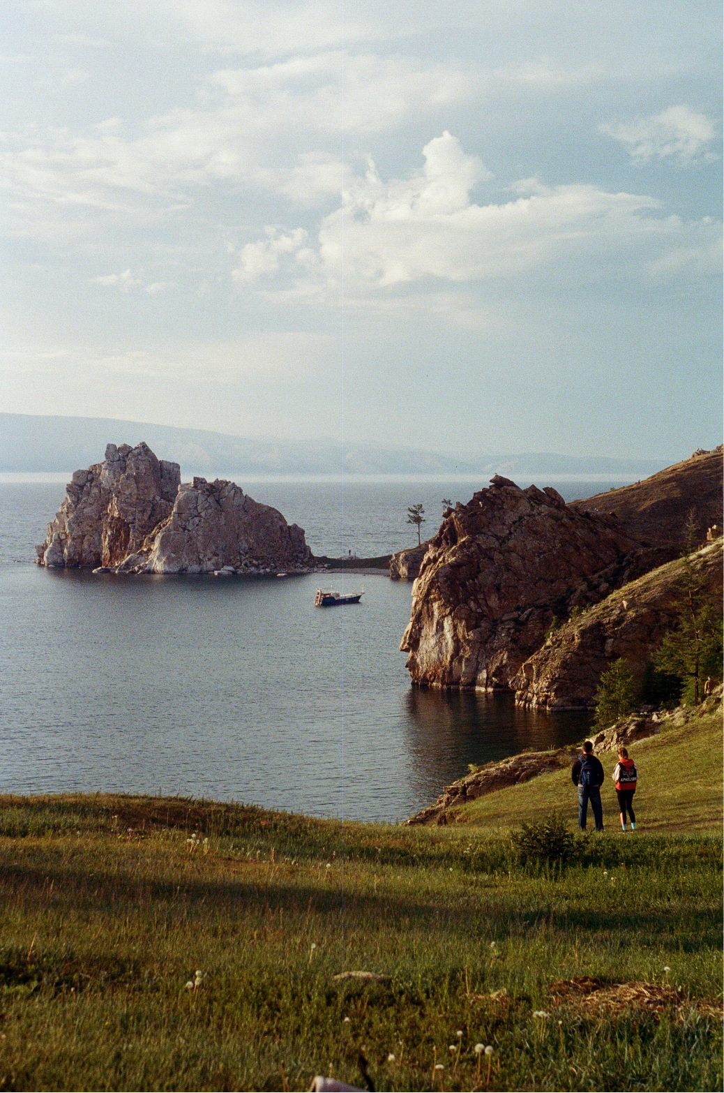 Green rolling hills in the forefront lead to a rocky cliff area by the sea. It extends into a little peninsula near the coast. A couple admires the sea on a grassy area to the right of the picture.