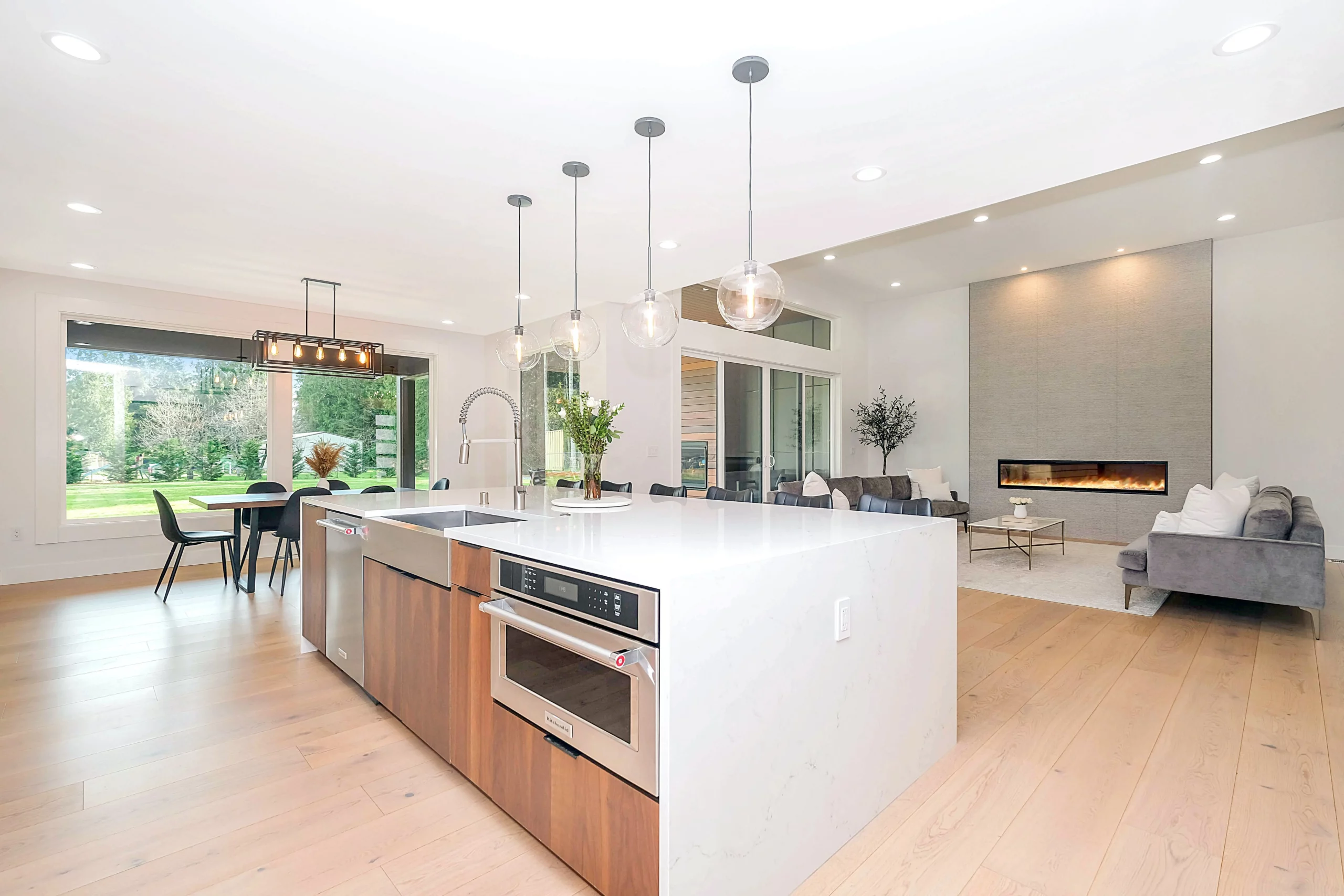 Cocina contemporánea con una gran isla con horno y fregadero, con la sala de estar al fondo y una gran ventana que da al jardín a la izquierda