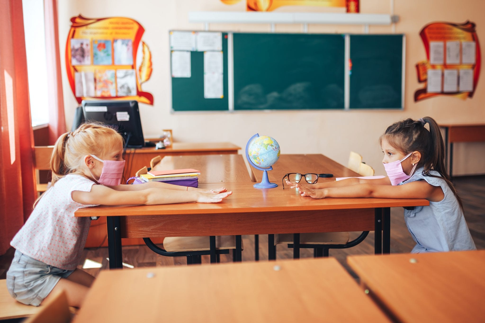 Two schoolgirls in medical masks are sitting at a school desk, opposite each other, group session, back to school, teaching children, social distance during epidemic. (Photo licensed to The Zebra Press by Adobe Stock)