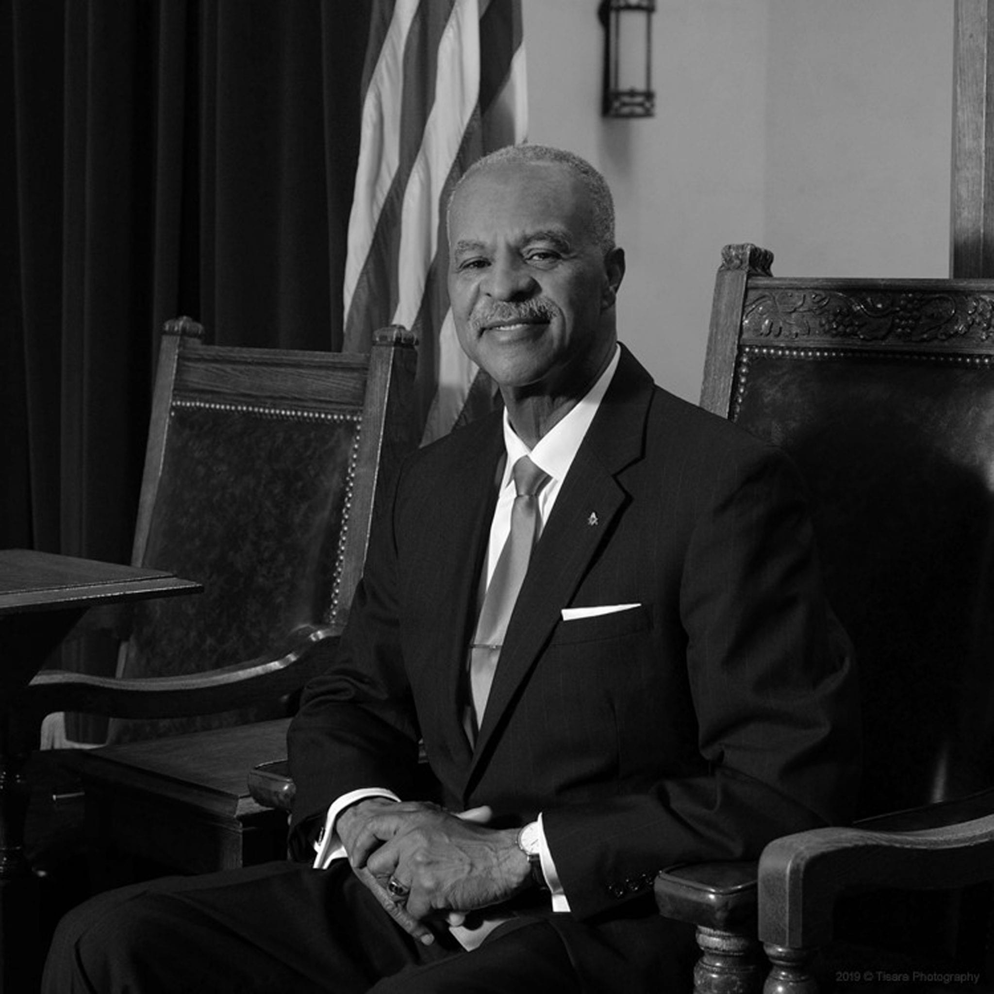 Black man in coat and tie sitting portrait syle in regal chair with flags nearby