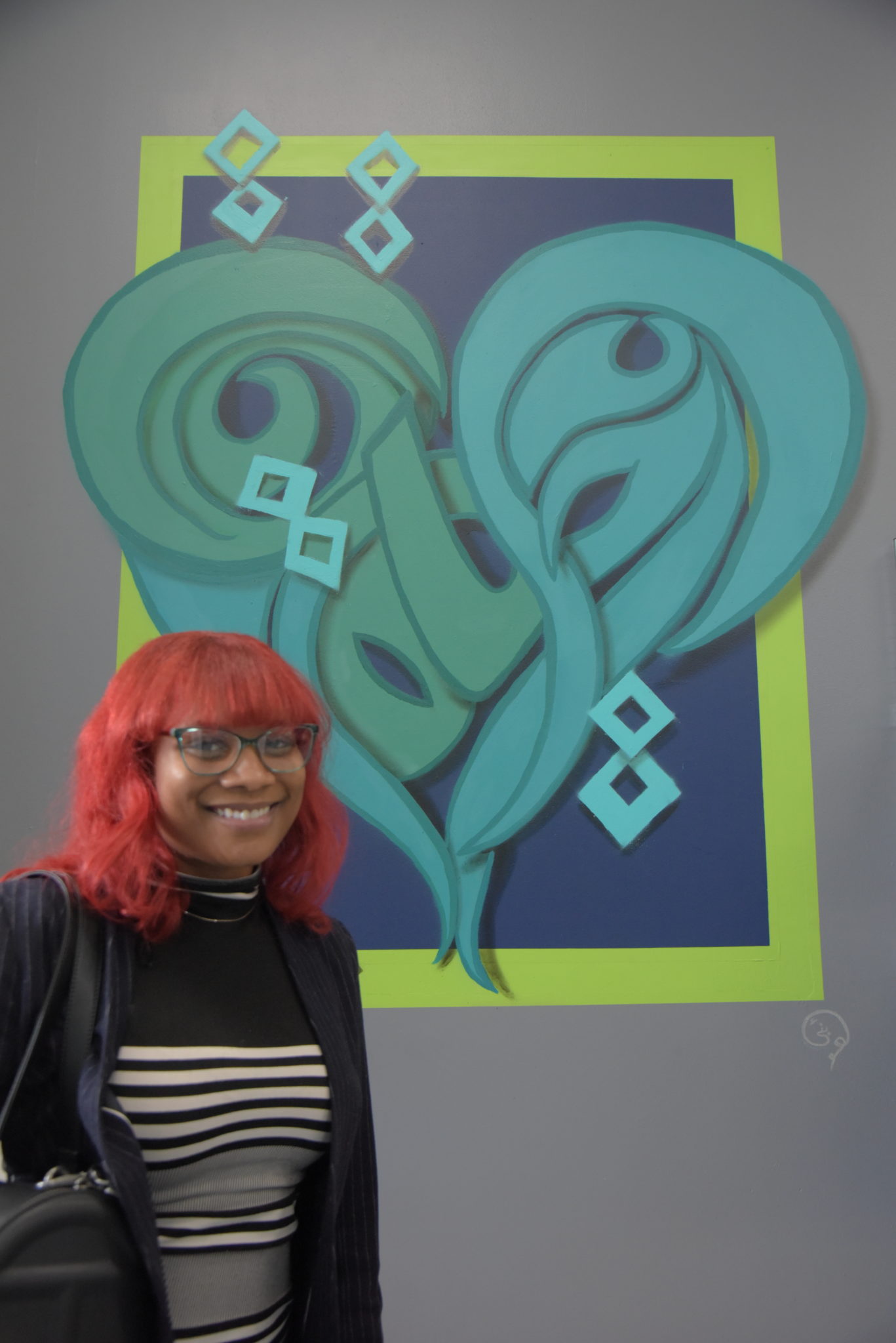 a woman with red hair and glasses poses in front of her mural of the arabic script for "alive" written in the form of a heart in turquoise