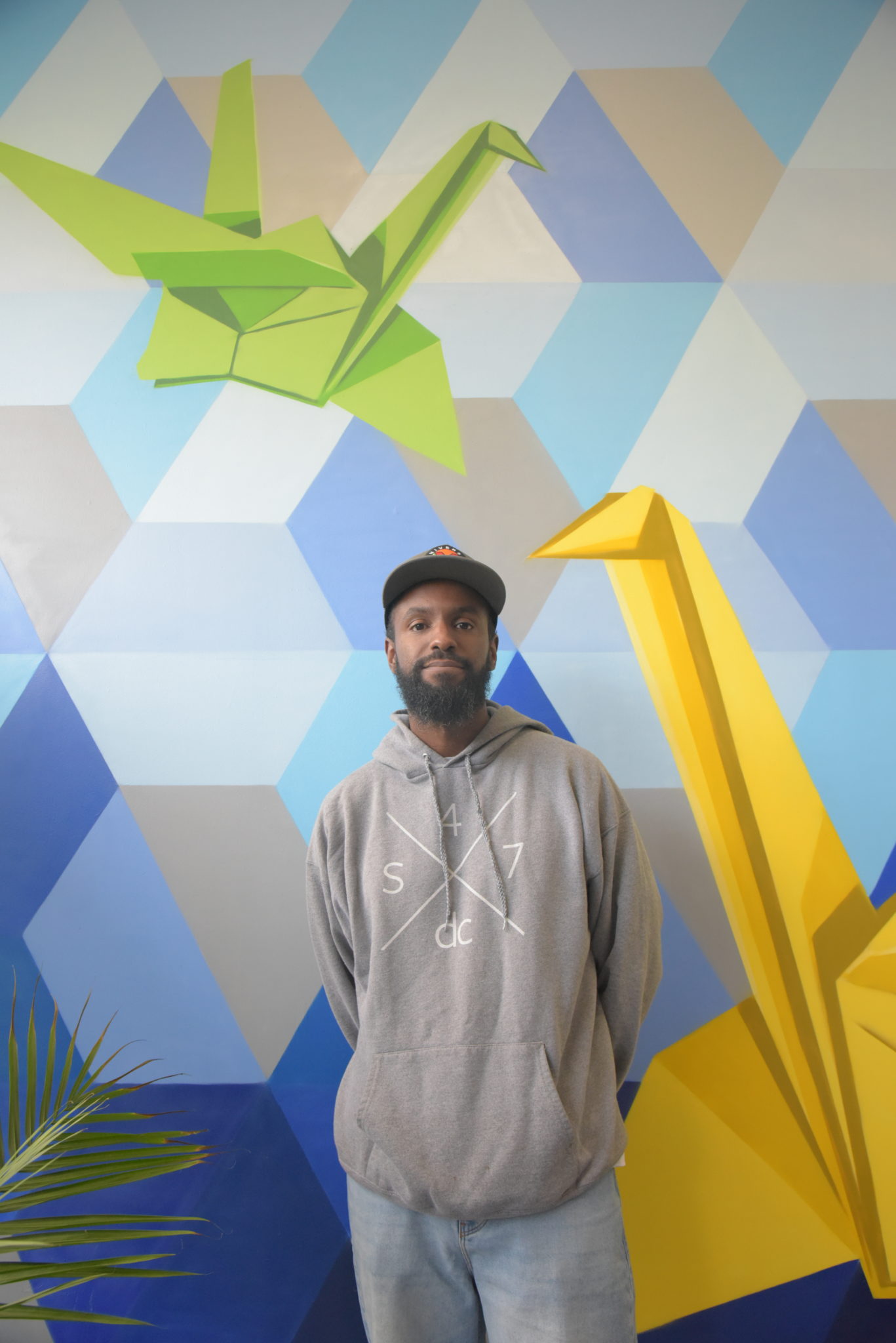 a man in a hat poses in front of a wall with a mural of two origami birds 