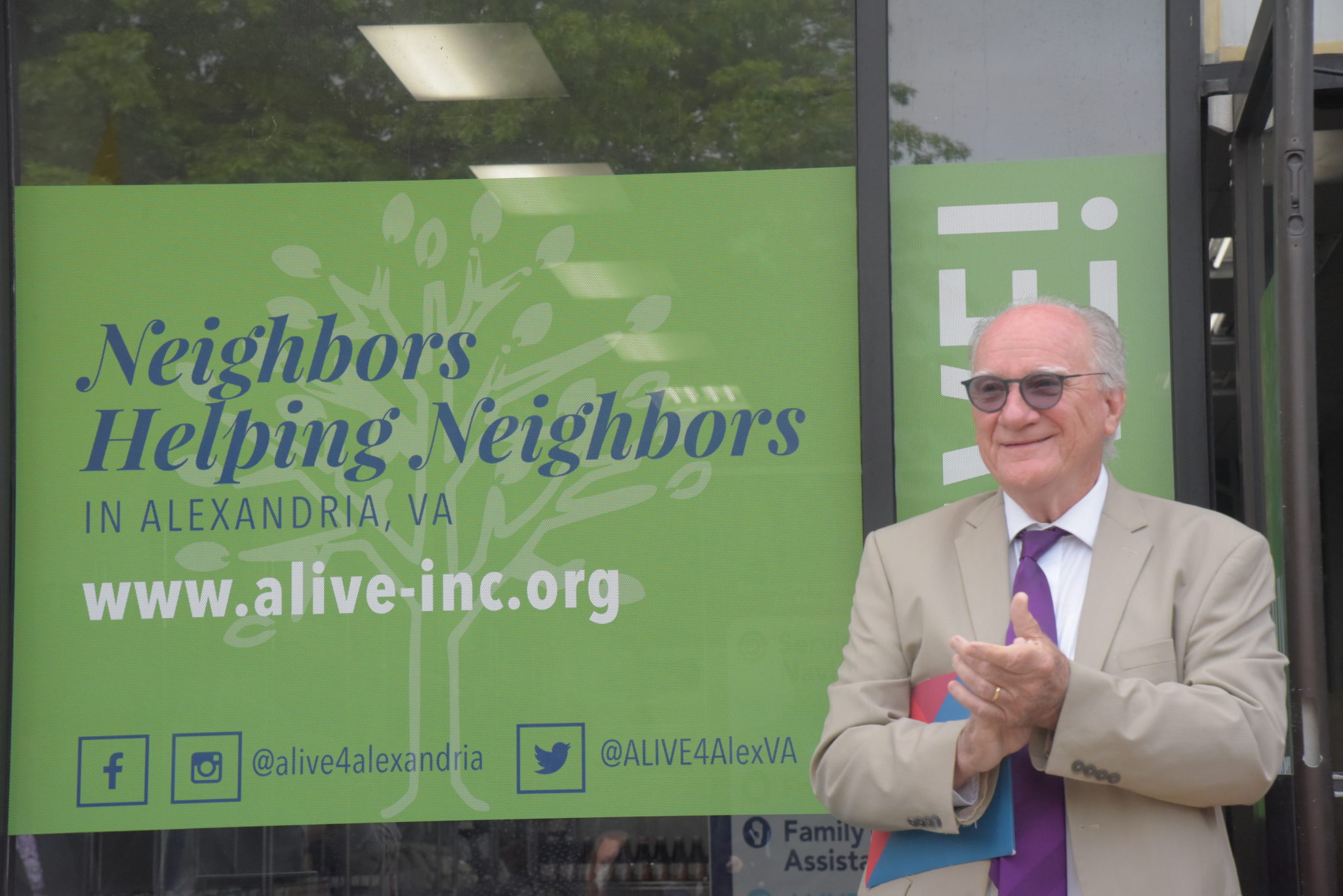 a man in a tan suit and purple tie claps his hands together in front of the ALIVE! Food Hub 