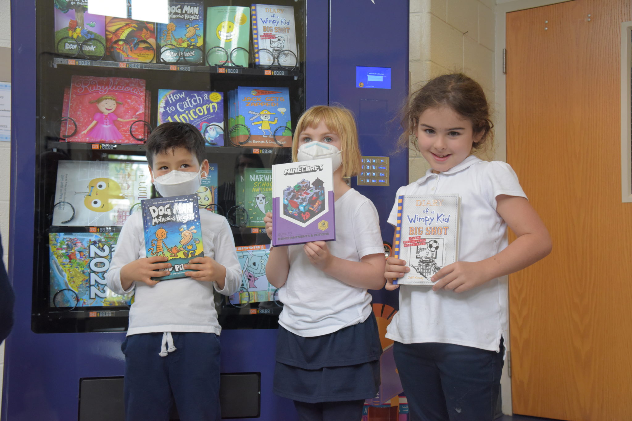 three school children stand in front of a book vending machine each with a book in their hands