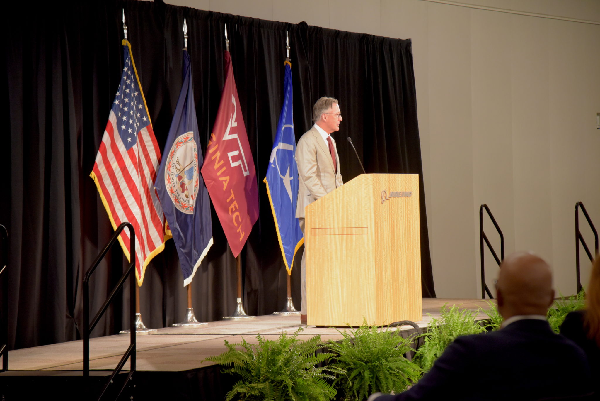 Virginia Tech President Tim Sands stands at podium with American Flag, Virginia Flag, Virginia Tech Flag, and Boeing Flags on stands behind him