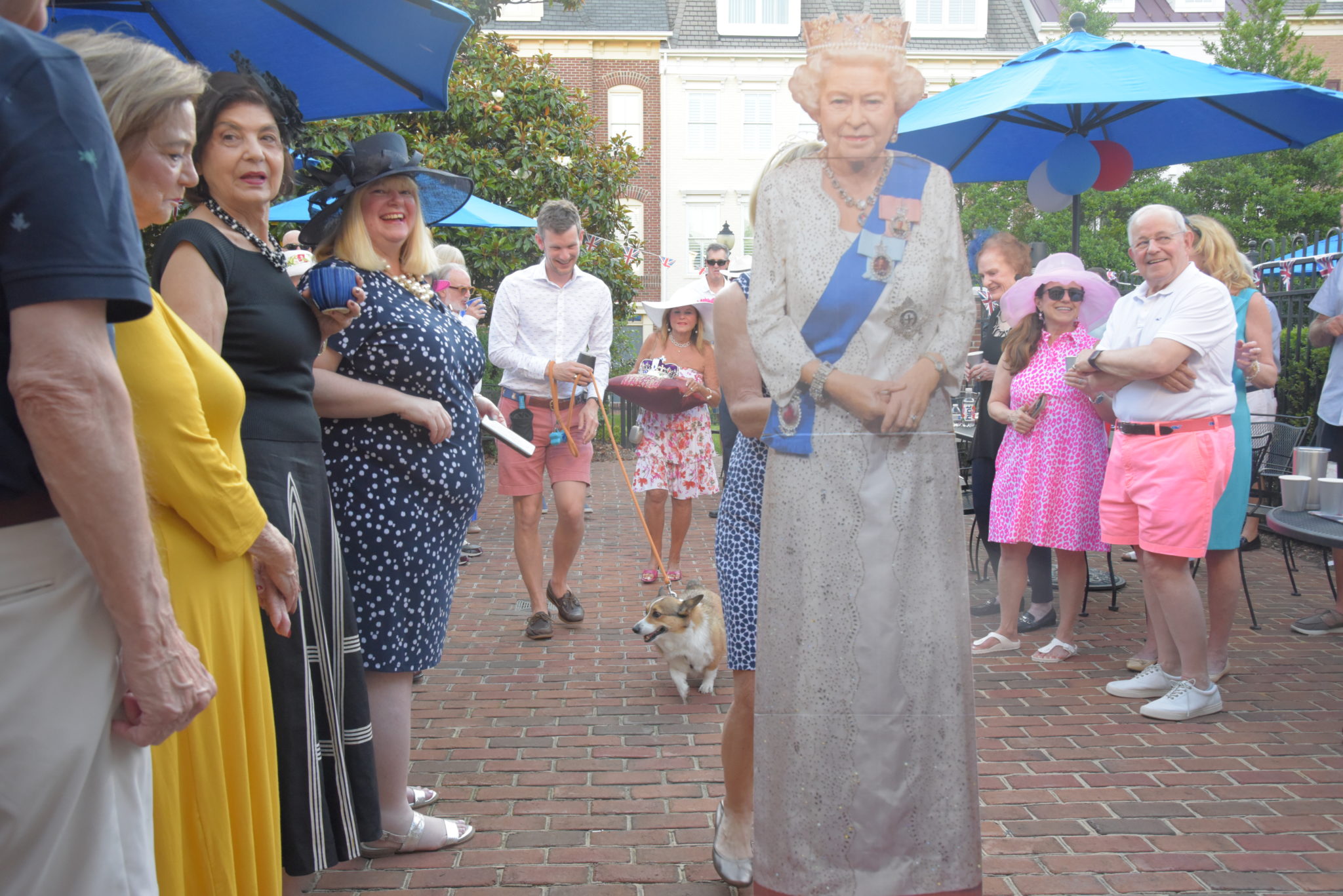 a crowd of people gathered on a patio around a cardboard cutout of Queen Elizabeth II