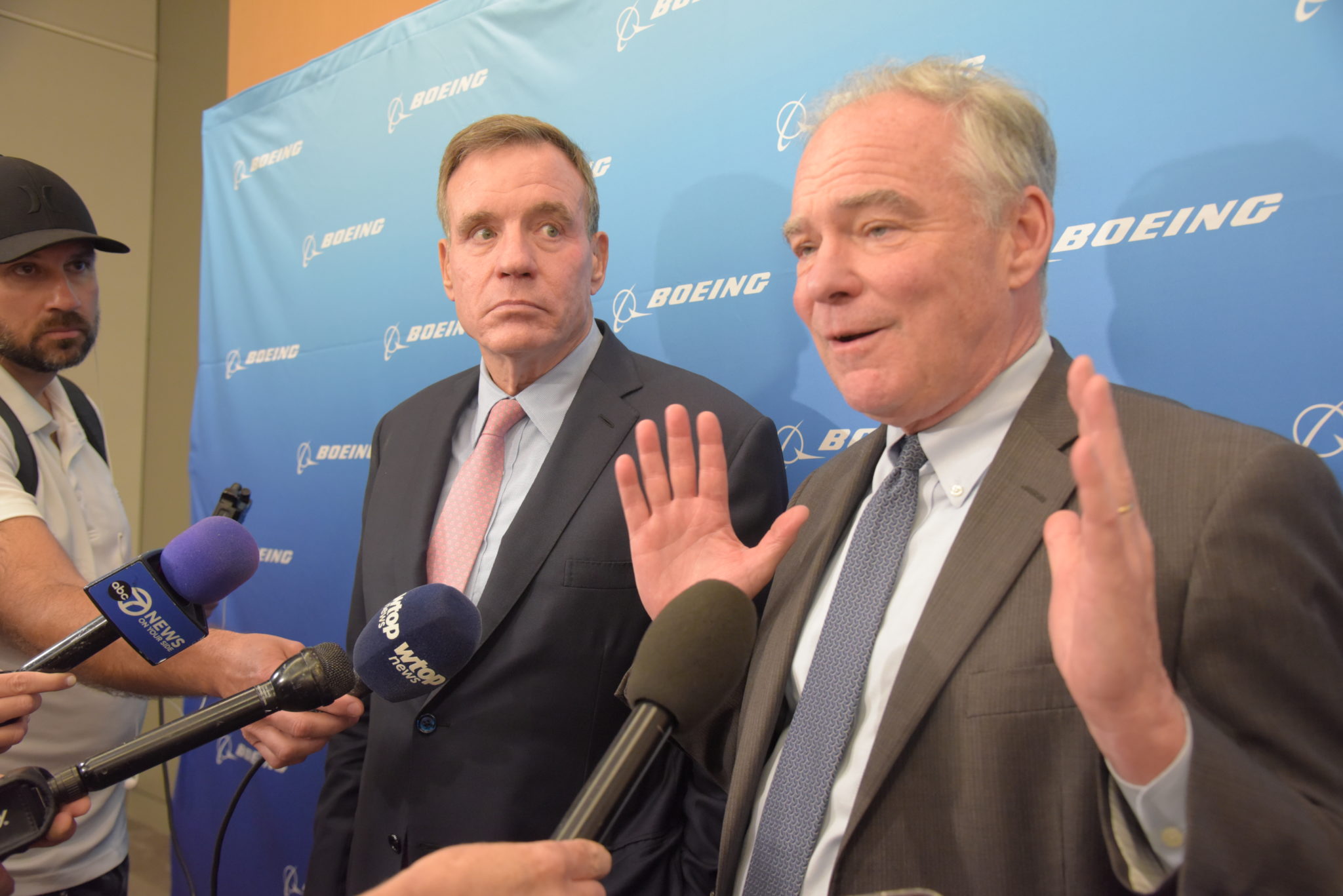 Senators Mark Warner and Tim Kaine stand in front of a Boeing sheet with cameras and microphones pointed in their faces, Senator Kaine raises his hands in front of him, palms out