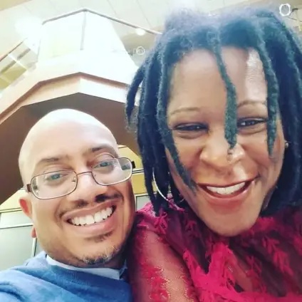 two African American people pose for a selfie, man on left wears glasses and a blue shirt, woman on right has short dreads and wears a red shirt