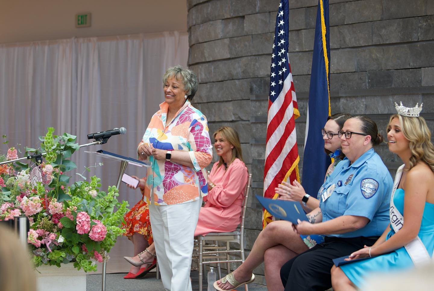 a woman in white pants and a pink shirt stands at a podium, a panel of people sit in chairs behind her