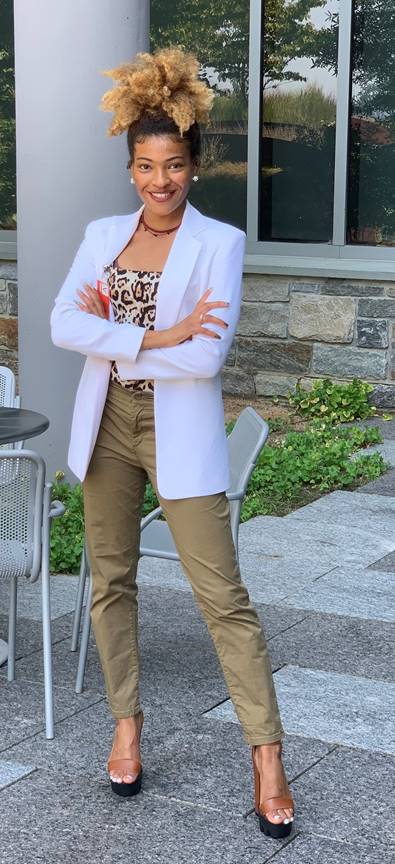 a woman stands in a white blazer, khaki pants, high heels, and a ponytail