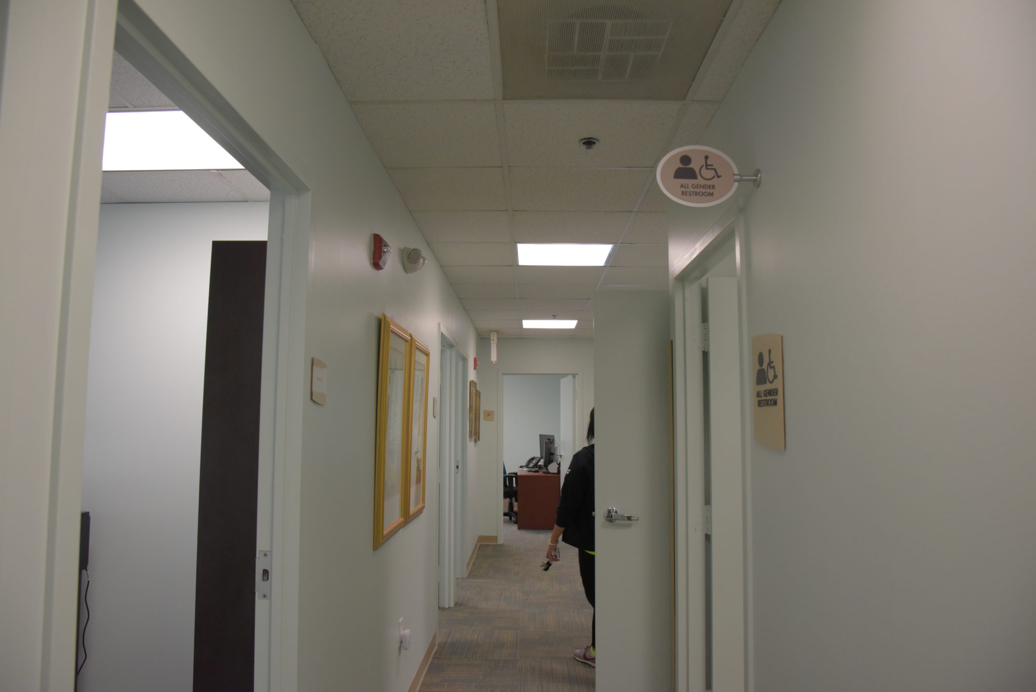 a hall way down a doctors office, a sign on a wall reads "all gender bathroom"