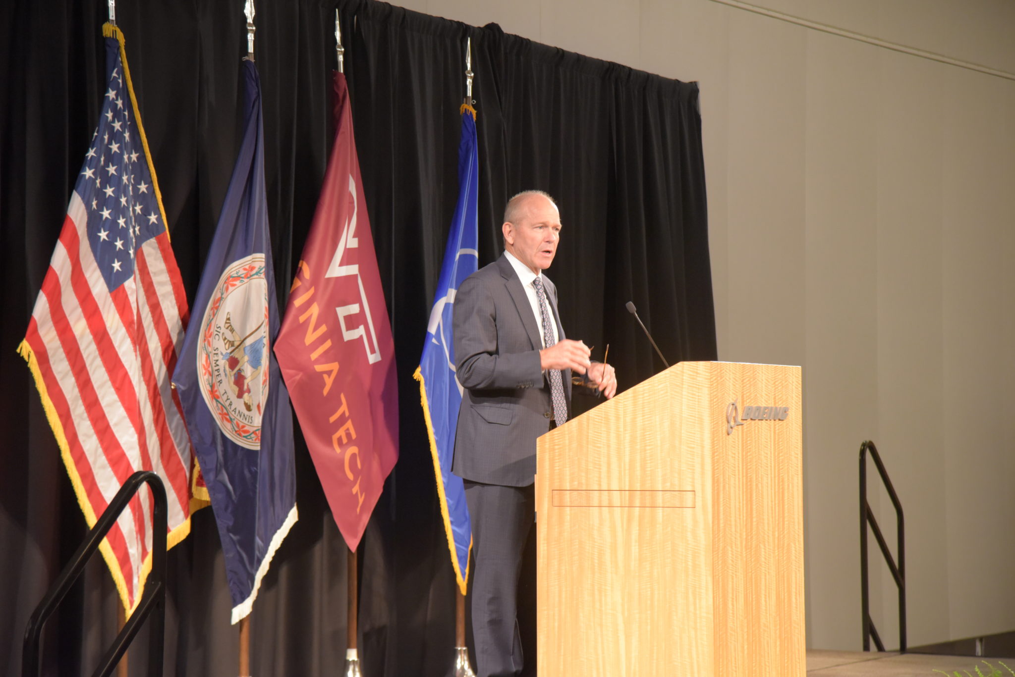 Boeing CEO Dave Calhoun stands at podium with American Flag, Virginia Flag, Virginia Tech Flag, and Boeing Flags on stands behind him