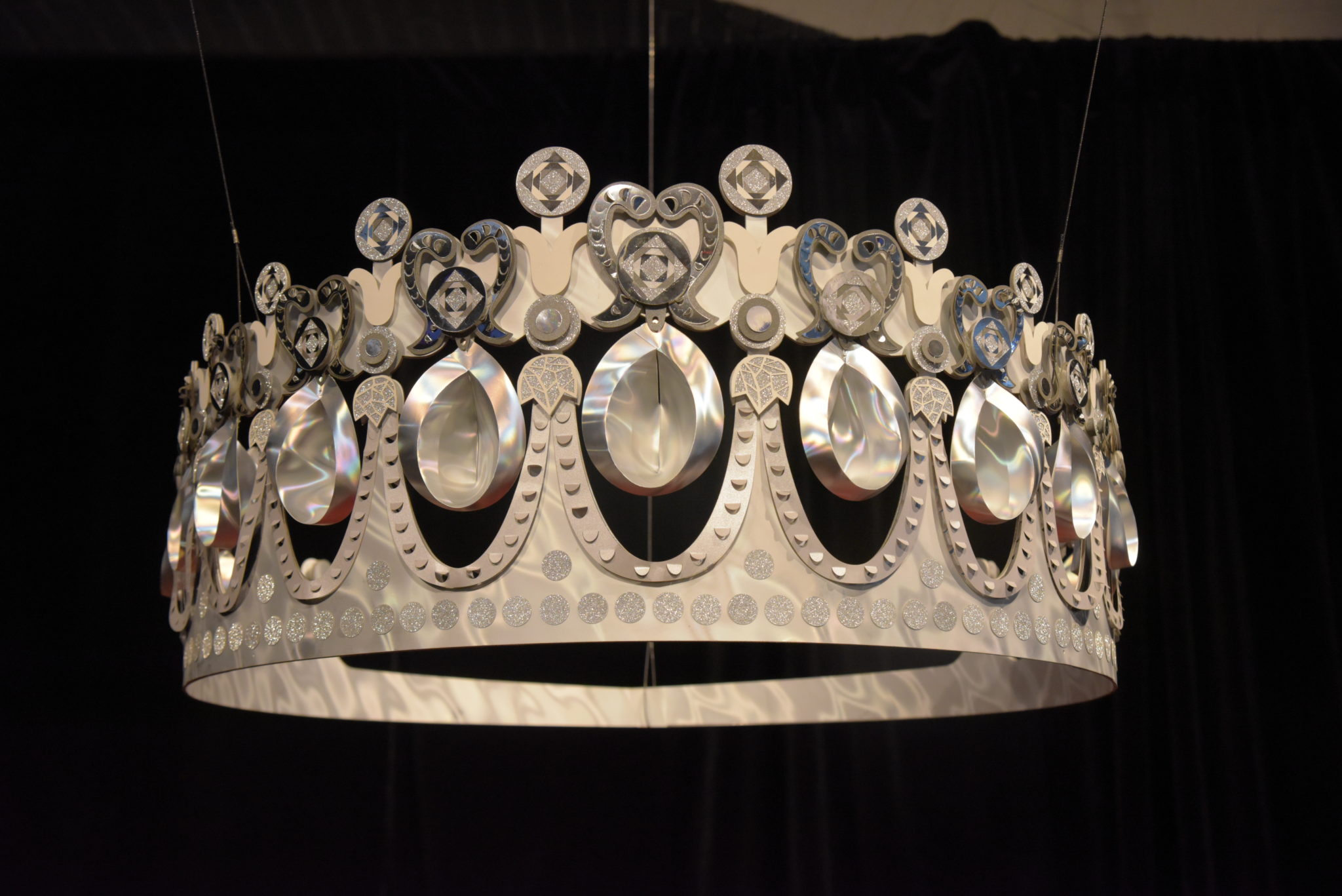 a paper sculpture of Princess Diana's tiara suspended from a ceiling, in front of a black curtain