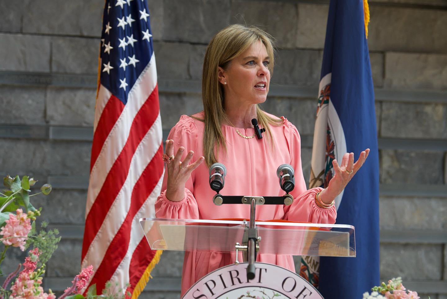 First Lady of Virginia Suzanne S. Youngkin wears pink dress, raises hands at a podium, flags of US and Virginia stand behind her