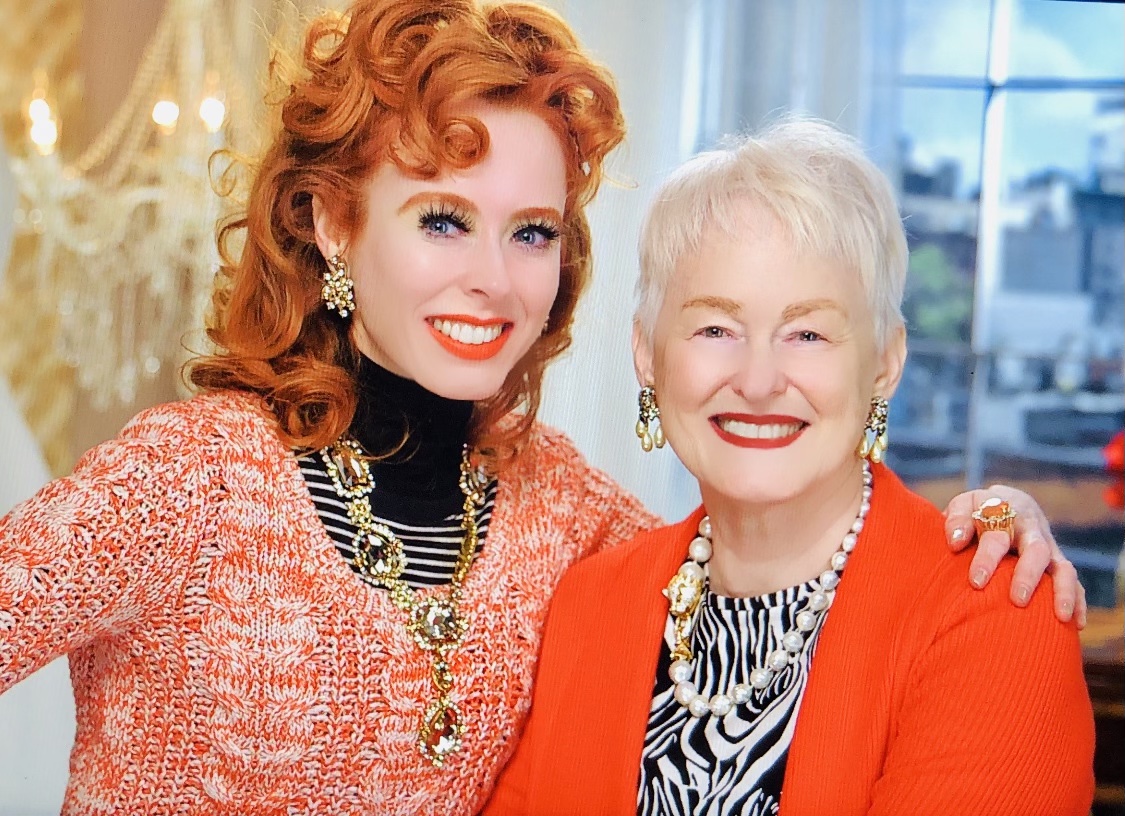 A red head woman stands next to her mother with white hair, both wear red sweaters, arms around one another