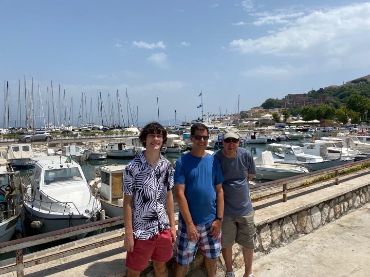 the men stand shoulder to shoulder in front of a bay filled with sail boats