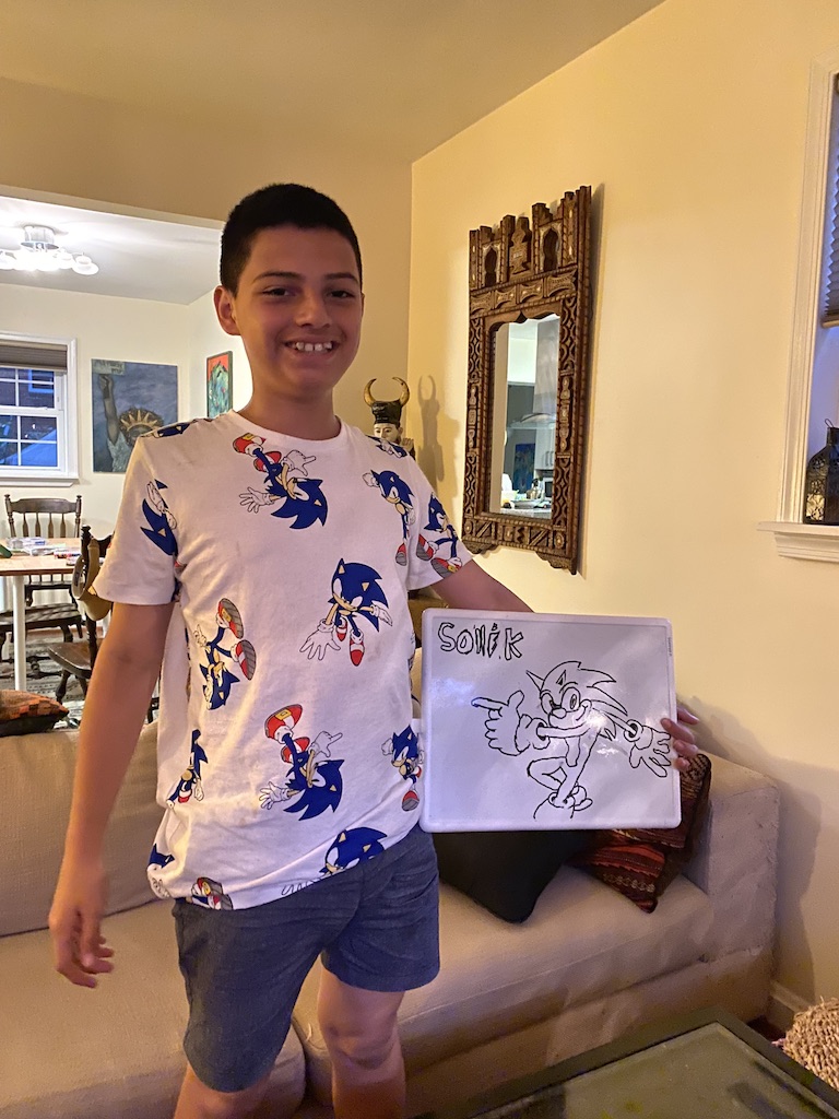 A boy holds up a sheet of paper with a hand drawn image of Sonic the Hedgehog