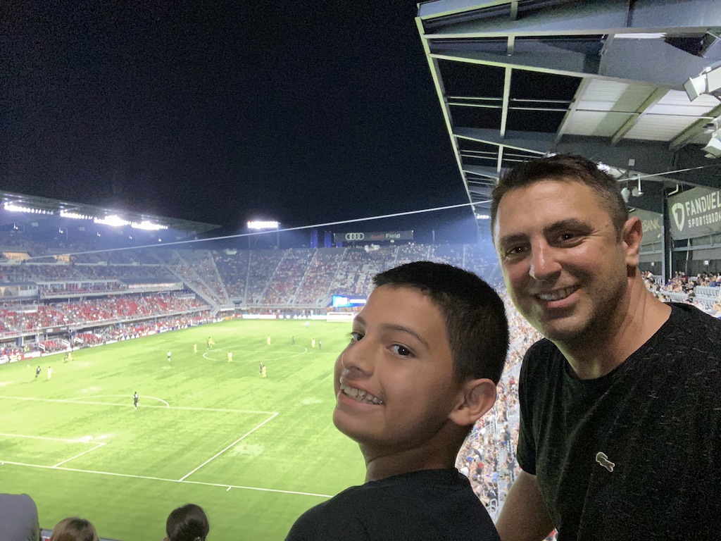 a young boy and his foster father smile at the camera while watching soccer at audi field