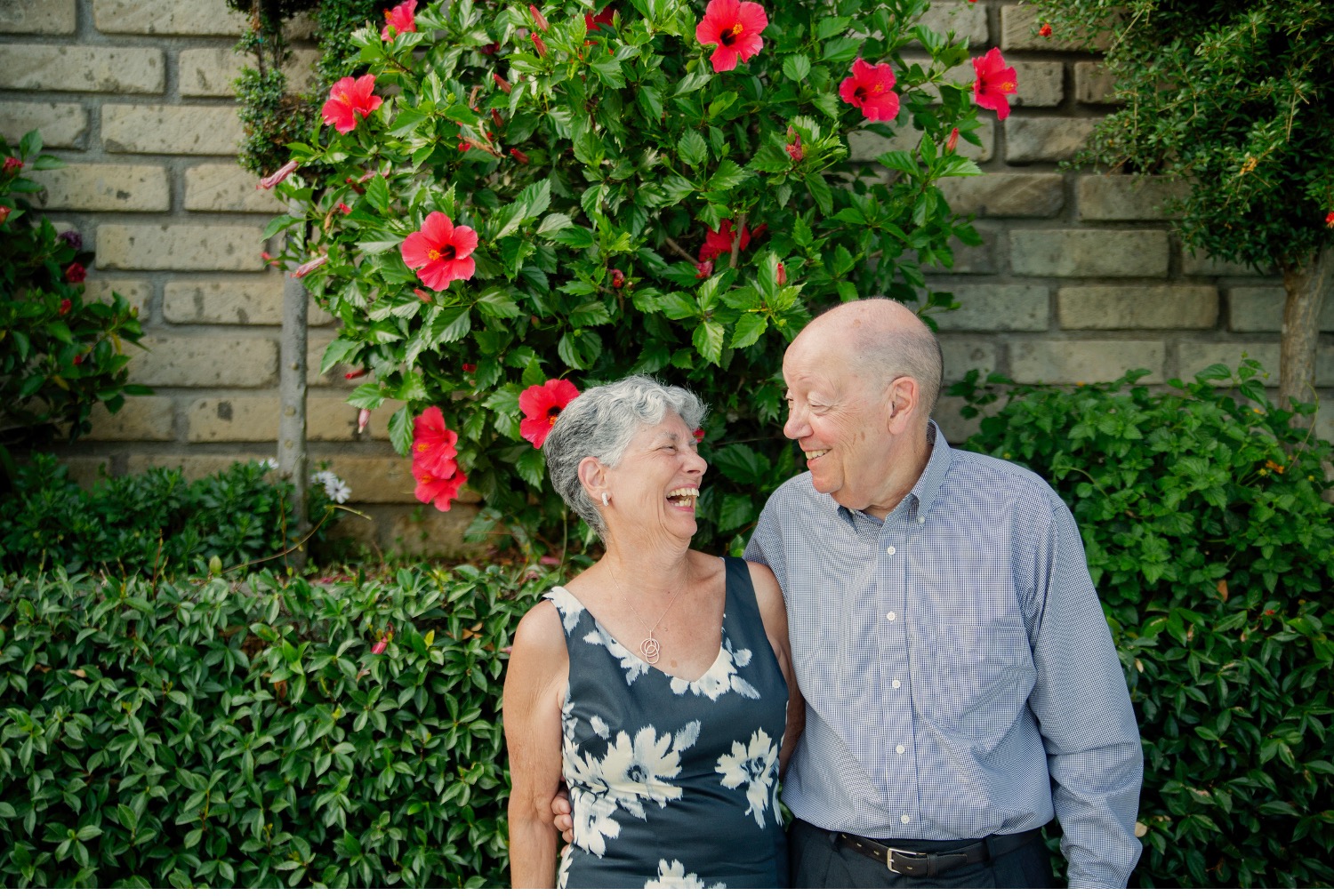 a man and woman stand in front of a wall covered with vines, smiling widely at each other