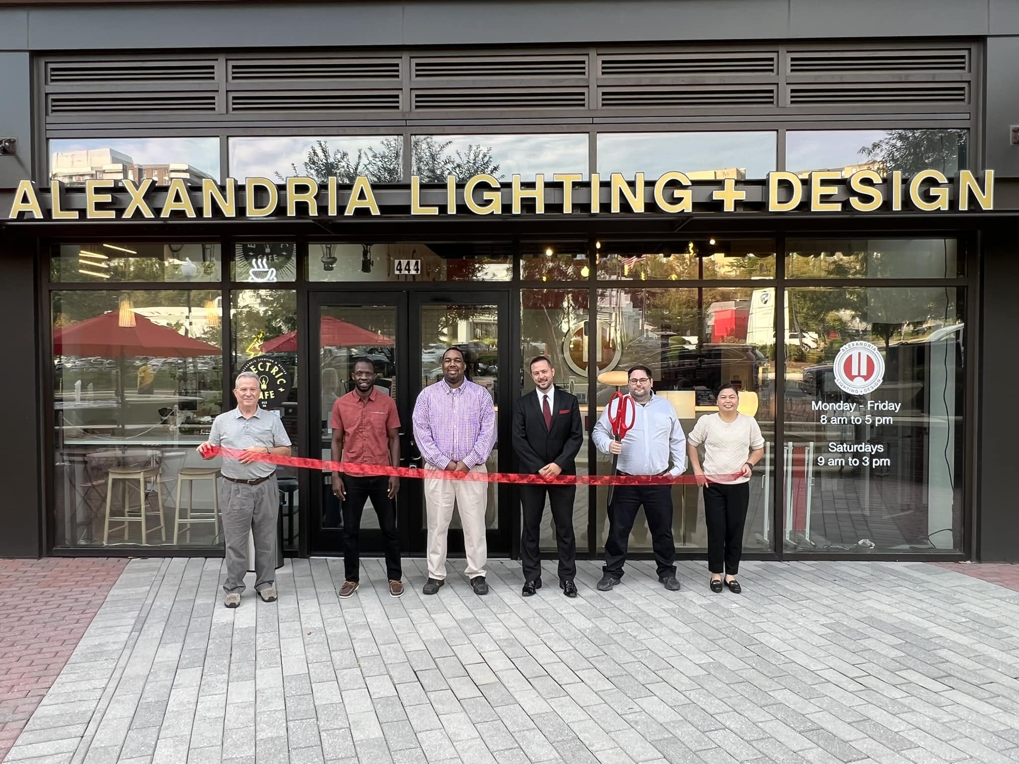 six people stand behind a ribbon with big scissors in front of the new Alexandria Lighting in Alexandria Virginia
