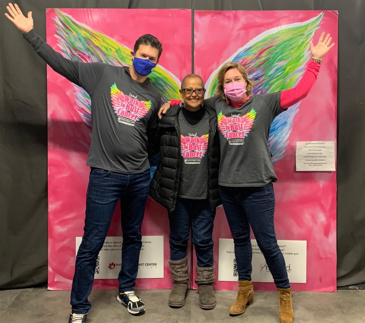 three people link arms in front of a mural of oversize angel wings