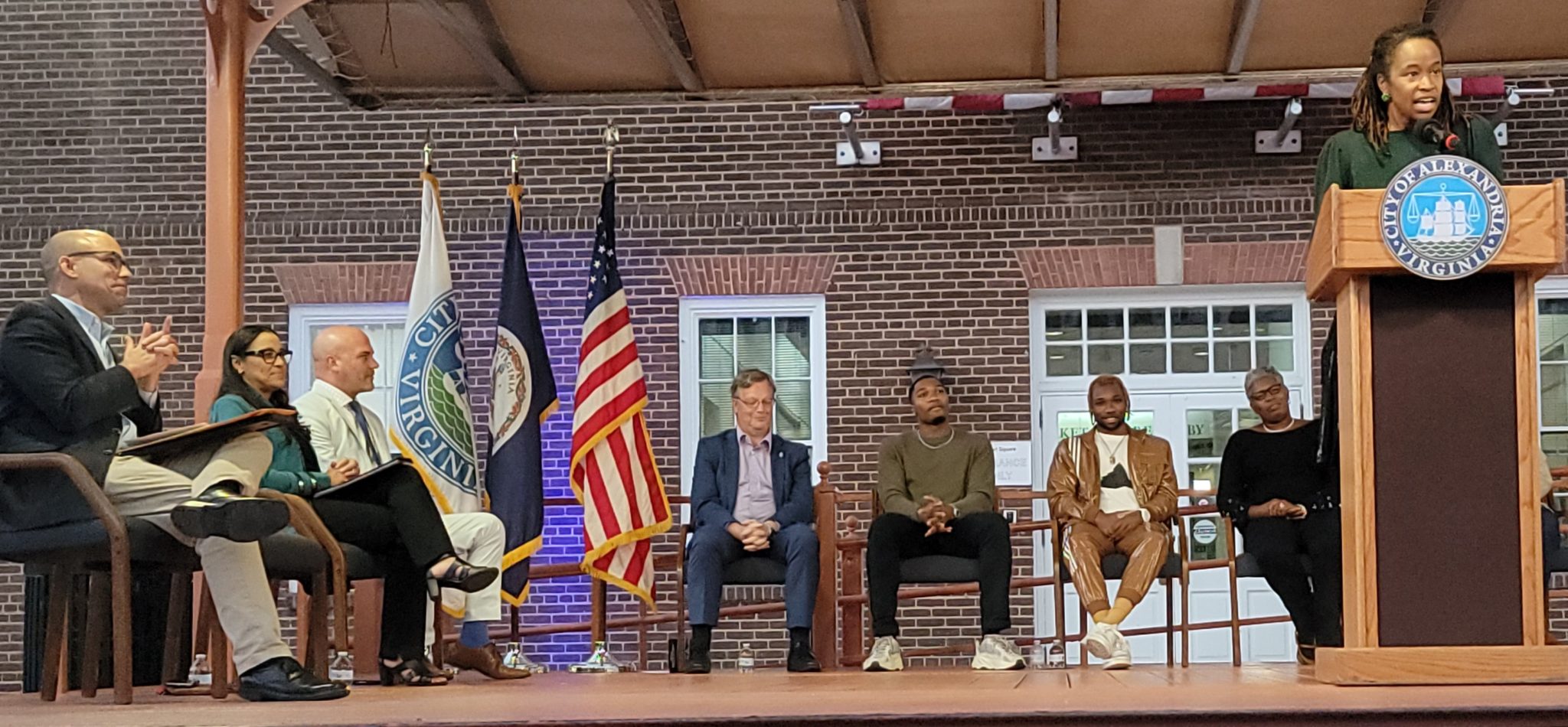 seven people sit in chairs on stage as woman speaks at podium