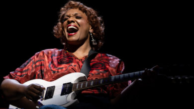 An African American woman wearing a red dress sits in a wheelchair center stage holding and playing a white electric guitar, singing with expression.