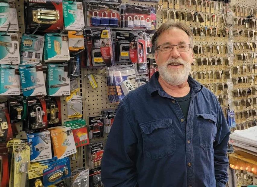 Man with beard smiling with hardware and keys hanging on wall behind him.