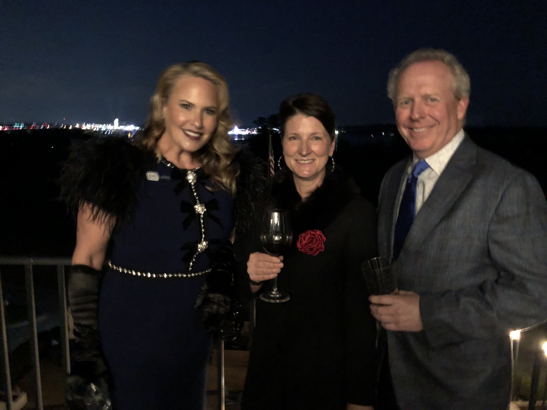 Three people at a night fundraising galla standing against a balcony railing with water and lights behind.