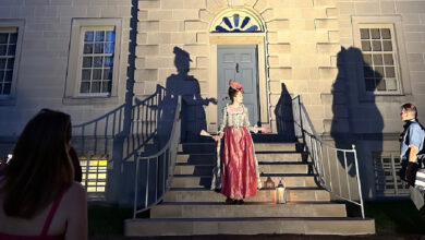 Woman in colonial dress stands at top of outside staired entryway of white building with blue arched door.