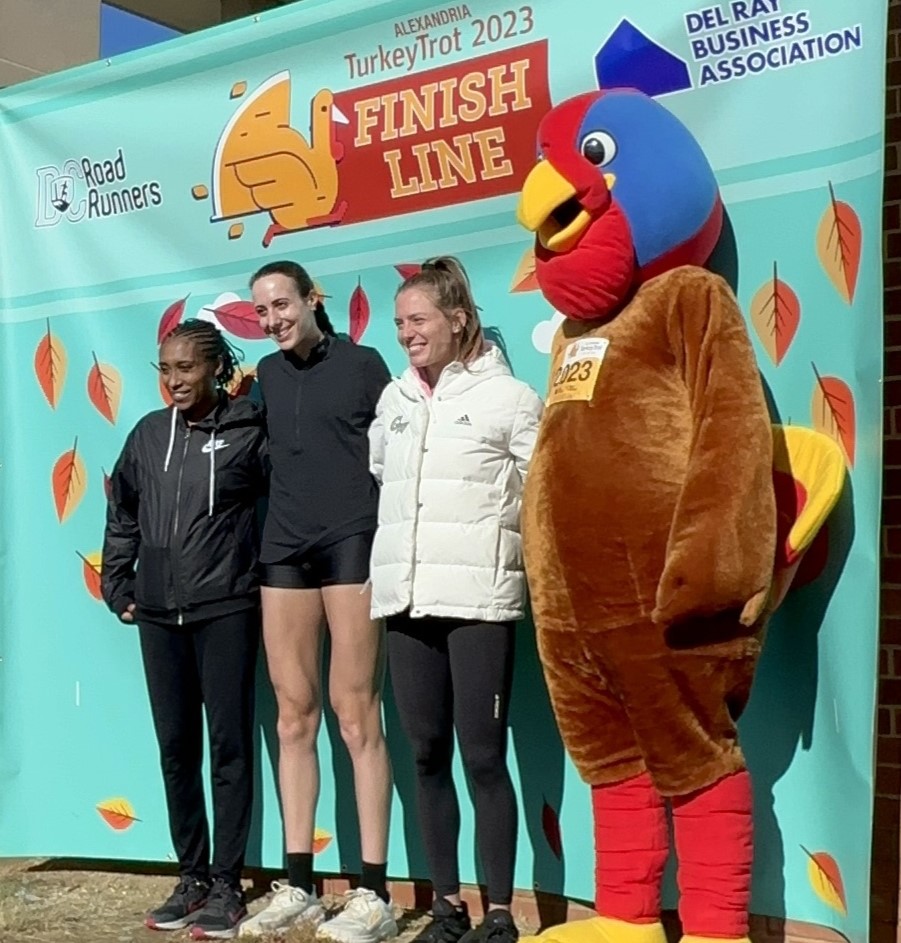 Three women standing with bug turkey mascot at finish line.