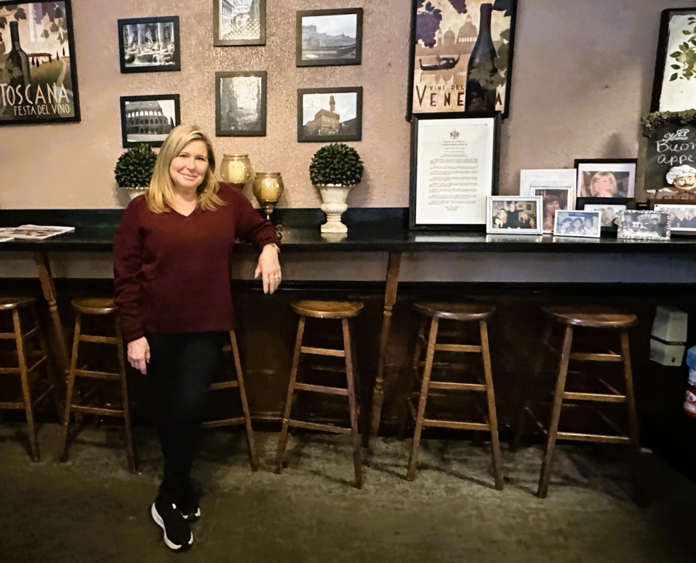 Caucasian blond lady leaning against bar facing camera with photos on wall behind her in Via Veneto restaurant