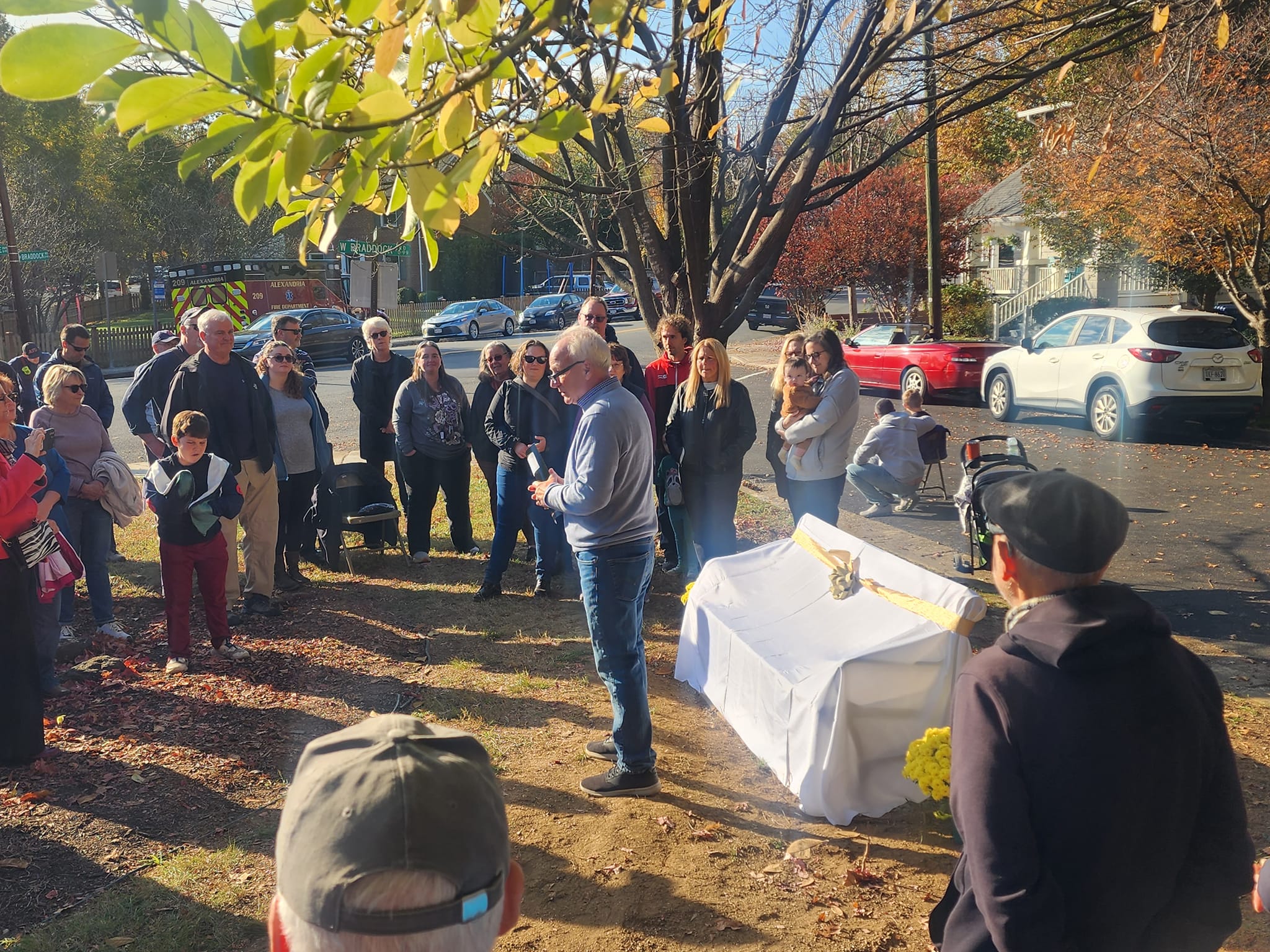 Crpwd gathers under large trees with a bench covered with a white sheet and bow.