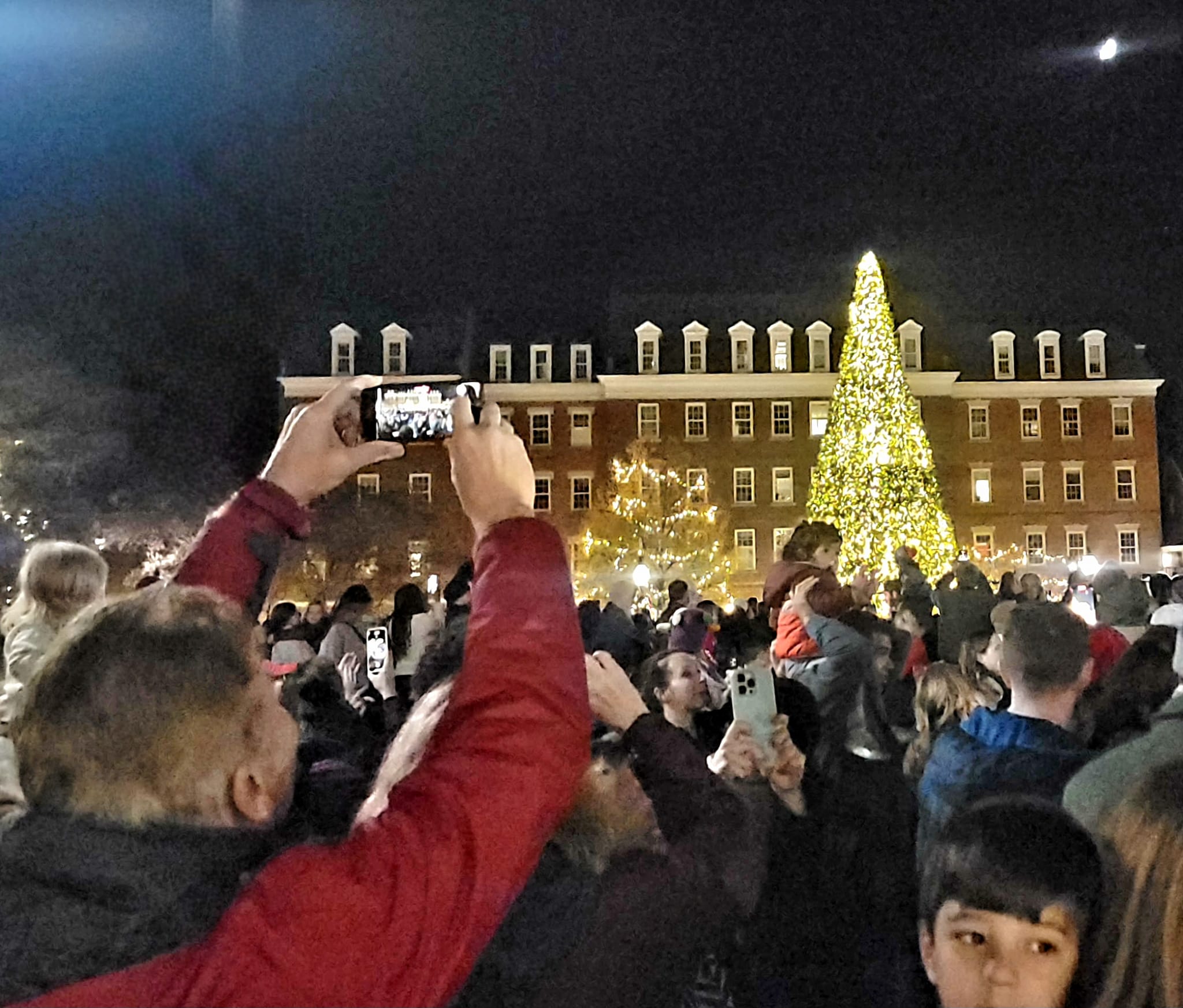 In foreground man is taking a photo at night of a family in front of large Christmas Tree in public square with hundreds of other people around.