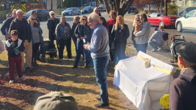 Crowd gathers under large trees with a bench covered with a white sheet and bow.