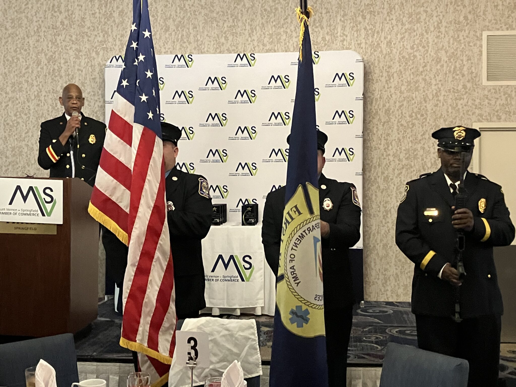 Men in uniforms holding flags at an indoor ceremony.