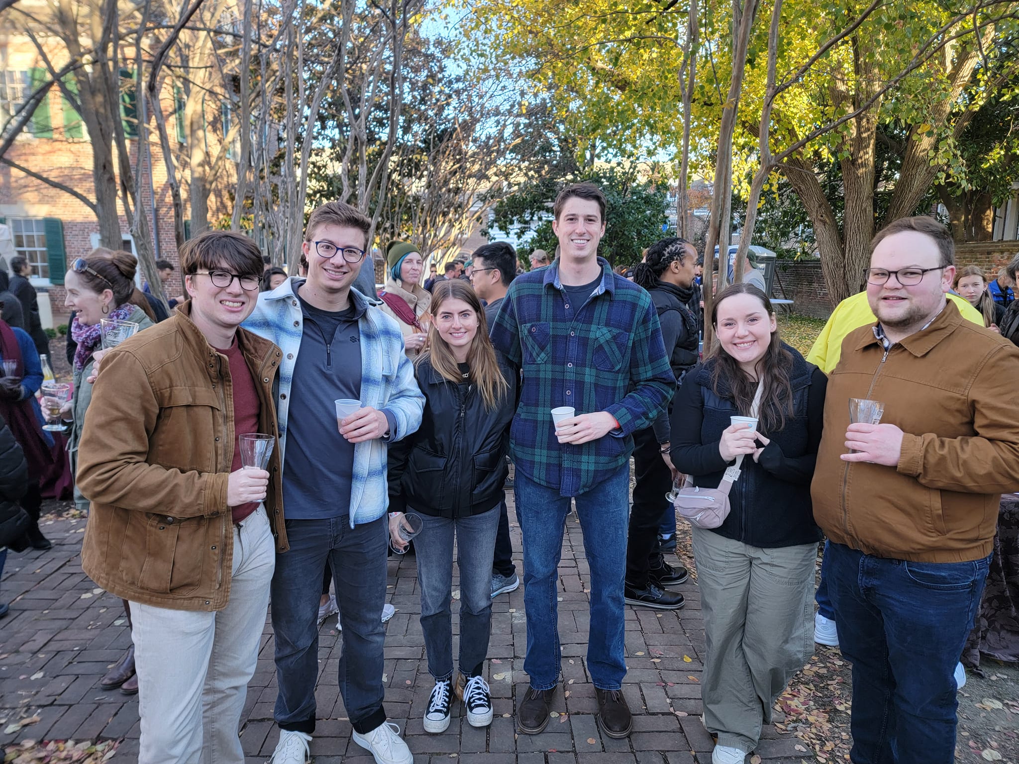 Seven people dressed in fall clothing holding cider glasses and smiling at camera.