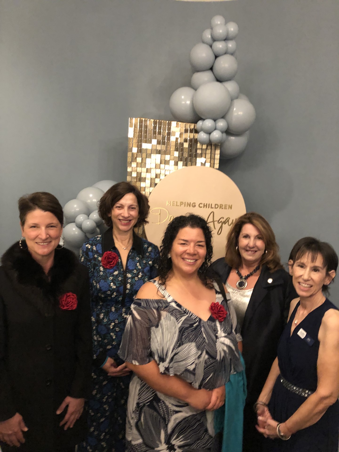 Five women standing in evening gowns in front of blue balloons.