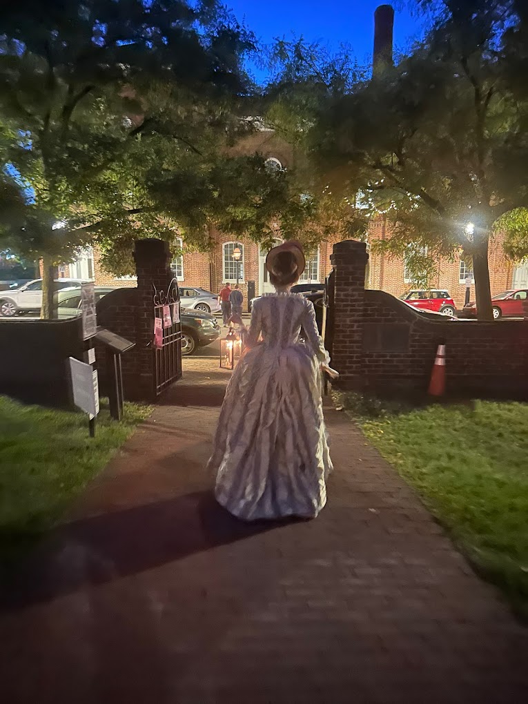 Woman dressed in colonial dress walks away on brick sidewalk toward brick gates.
