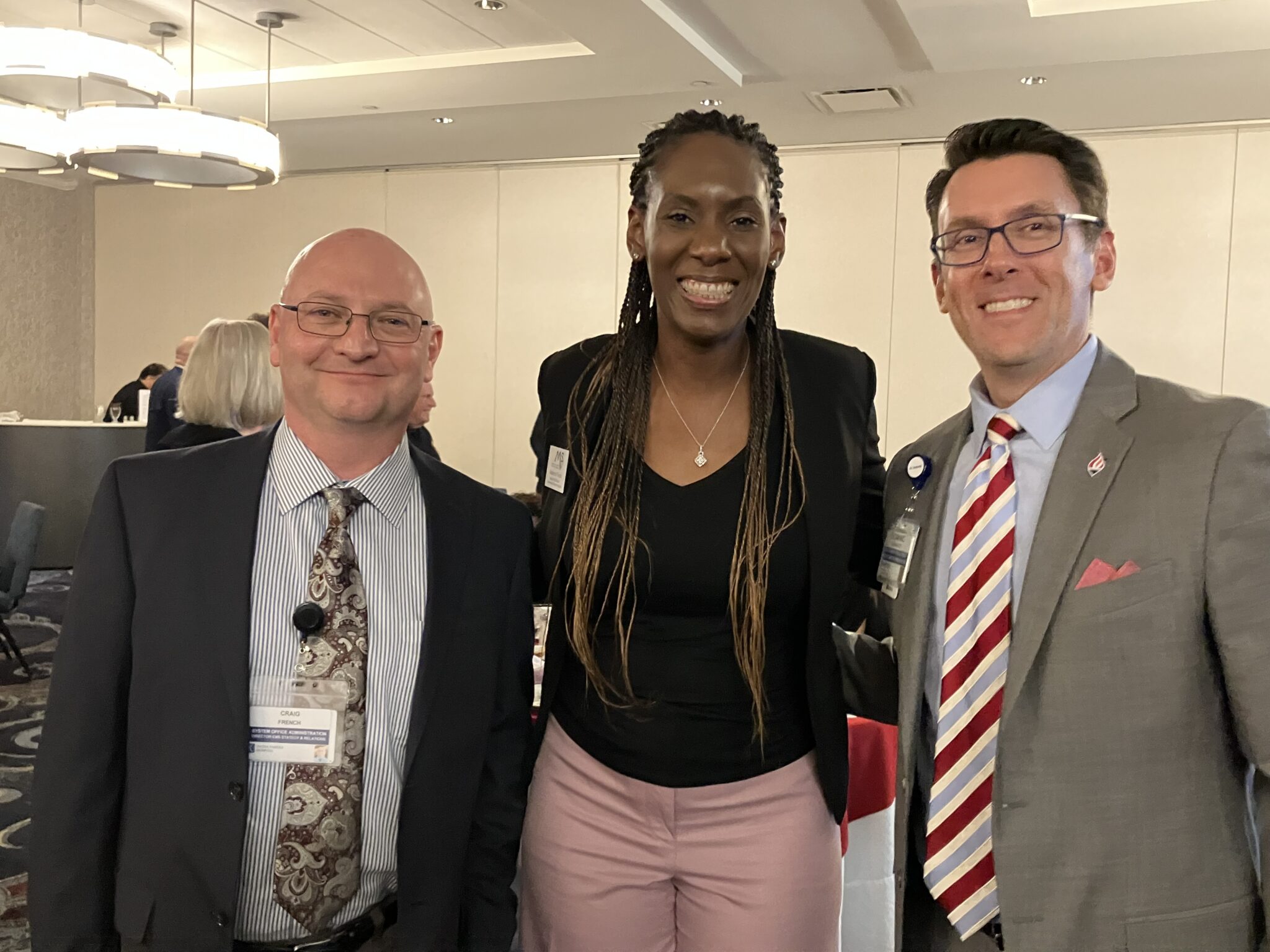 Three people looking at camera, bald white man in coat and tie on left, tall beautiful black woman in center, and middle-aged white man on right in coat and tie.