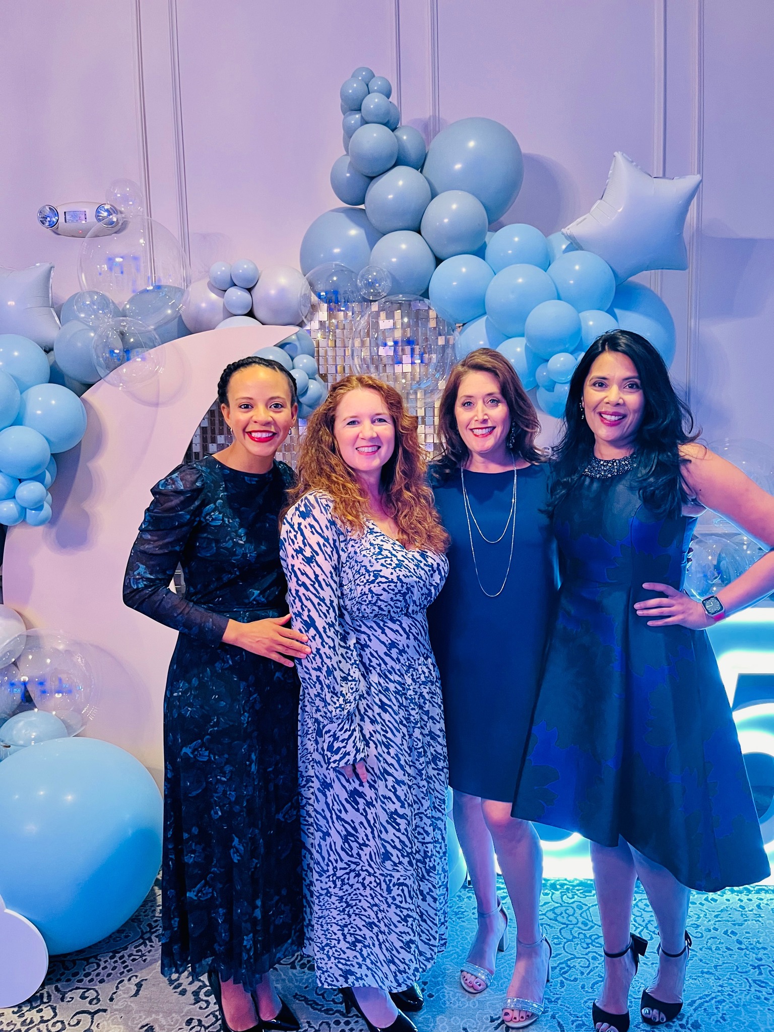 Four women dressed in blue evening gowns in front of a massive display of blue balloons.