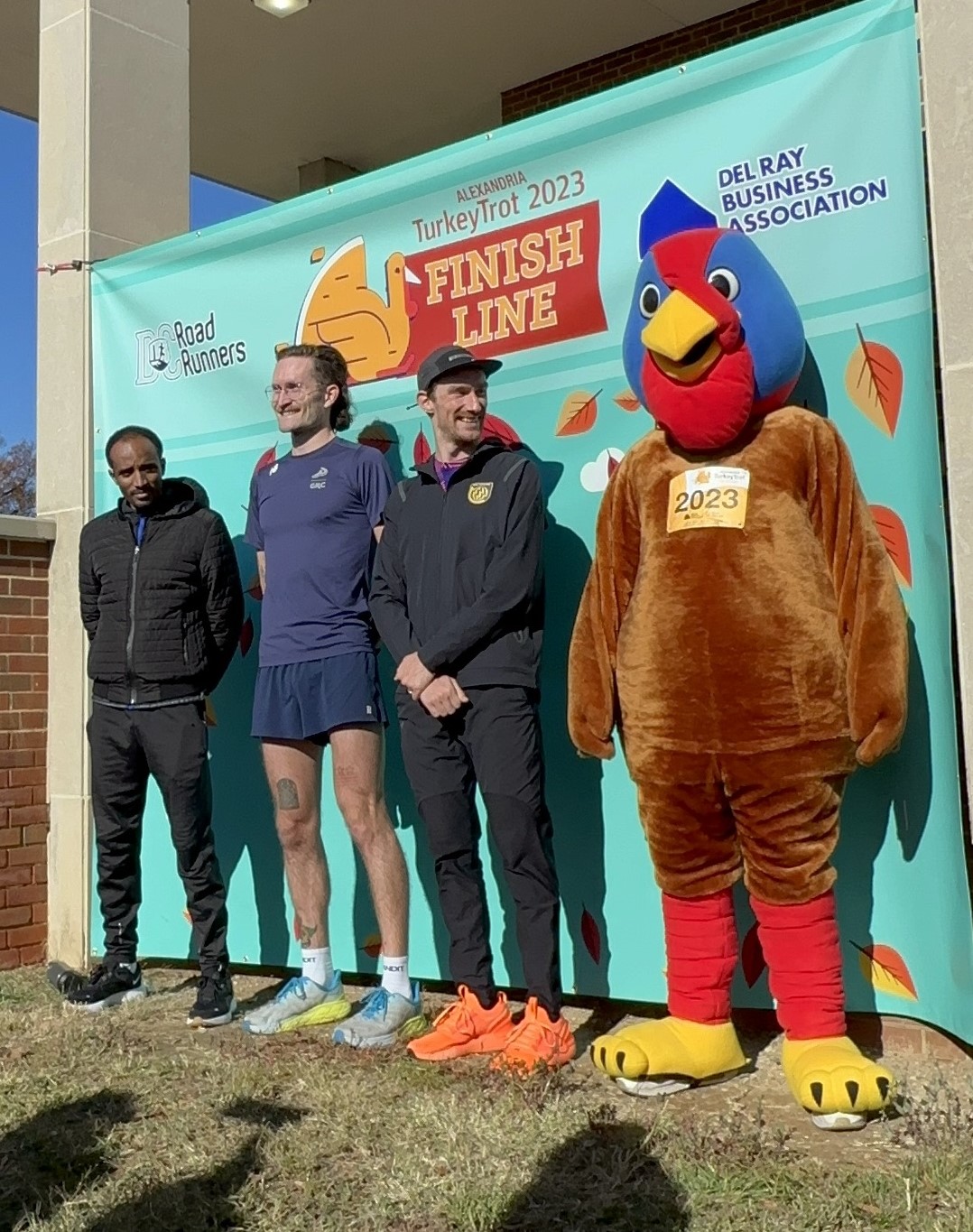 Three people and a big turkey mascot stand in front of a green finish line banner.
