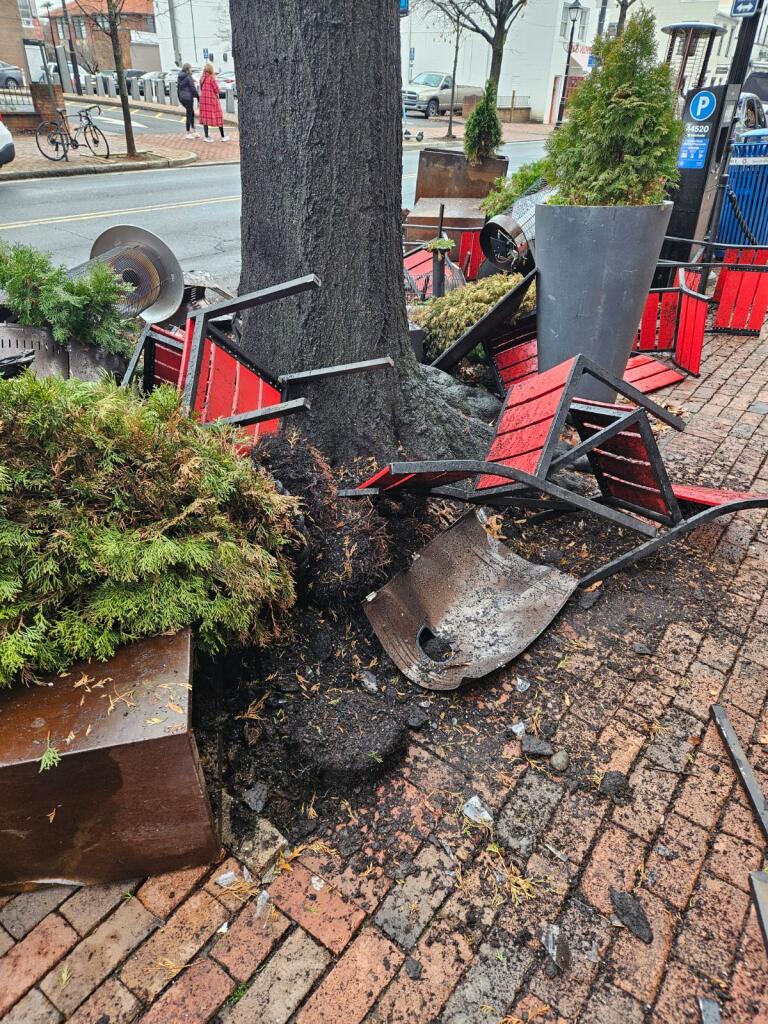 Chairs run over and mangled all over the sidewalk wedged against tree.